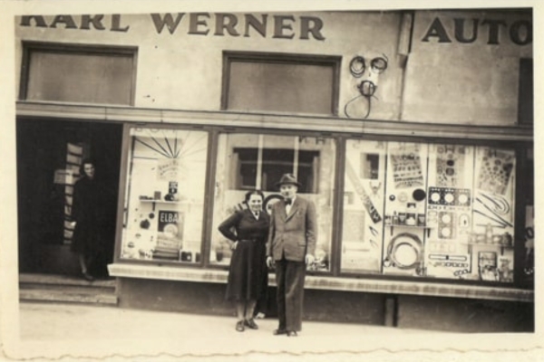 Ein altes Schwarz-Weiß-Foto zeigt einen Mann und eine Frau, die vor einem Geschäft mit der Aufschrift „Karl Werner Auto“ posieren. Im Schaufenster sind verschiedene Automobilartikel ausgestellt. Eine Person steht im Türrahmen.