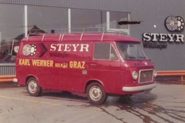 Ein roter Oldtimer-Transporter mit den Aufschriften „STEYR Walzlager“ und „KARL WERNER GRAZ“ an der Seite parkt in der Nähe eines Gebäudes mit „STEYR-Wälzlager“-Schild. Die Szenerie wirkt wie aus einer vergangenen Zeit, was sich am Fahrzeugdesign und der Bildqualität ablesen lässt.
