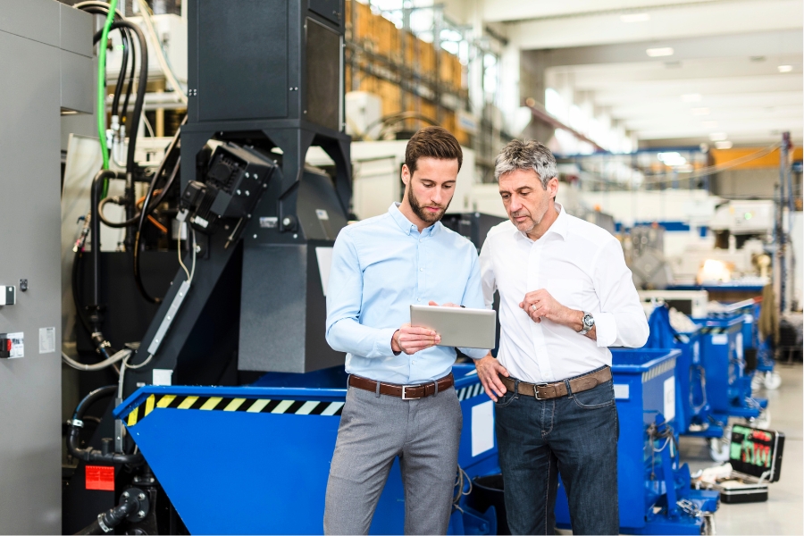 Zwei Männer in Businesskleidung in einer Produktionshalle. Beide schauen auf ein Tablet.