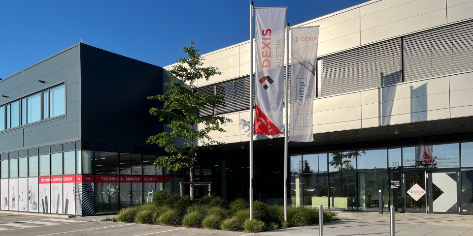 Modernes Industriegebäude mit grauer Fassade. Davor ist eine Grünflächen mit Baum angelegt, wo auch zwei Fahnen stehen mit "DEXIS Austria" und "multiCrimp".  Glaswände geben den Blick auf das Technik und Service Center sowie den Eingangsbereich des Bürogebäudes frei. Es ist strahlend blauer Himmel zu sehen.