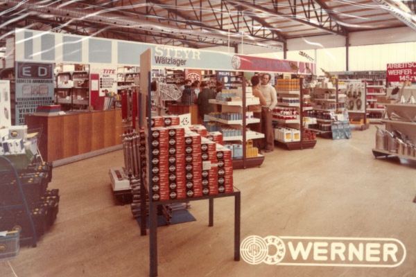 Ein historisches Foto vom Shop in Graz mit Regalen, in denen verschiedene Werkzeuge und Produkte ausgestellt sind. Eine Verkaufstheke ist sichtbar, darüber befinden sich Schilder und Werbung. Zwei Personen stöbern im Laden. Ein weißes „Werner“-Logo befindet sich im rechten Eck des Bildes.
