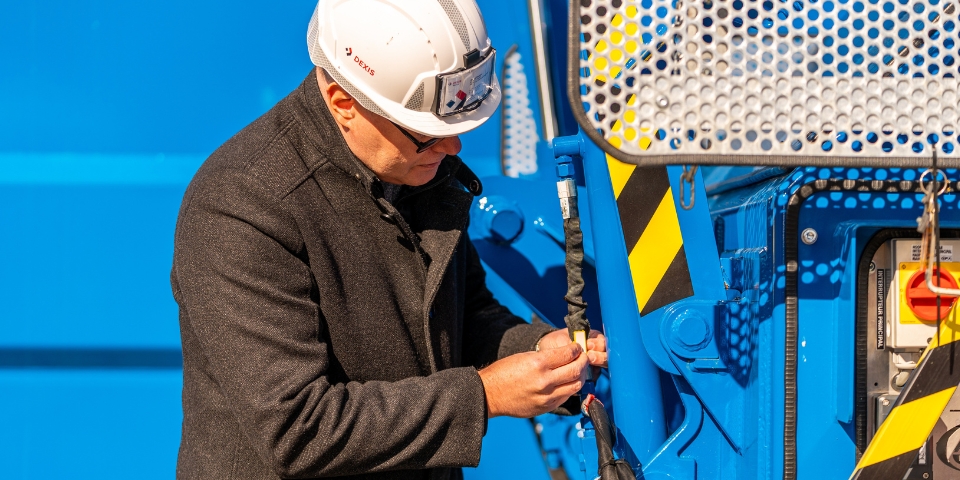 Eine Person mit weißem Schutzhelm, Brille und dunklem Mantel inspiziert eine  blaue Maschine. Die Person konzentriert sich auf einen Schlauch und ist von Industrieanlagen und einem schützenden Metallgitter umgeben.