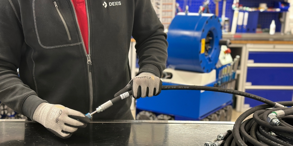 Ein Mann mit Handschuhen und dunkler Jacke arbeitet mit einem Schlauch auf einem schwarzen Tisch. Aufgerollte Schläuche mit Metallverbindern sind sichtbar, im Hintergrund ist eine blaue Hydraulikschlauchpresse zu sehen.