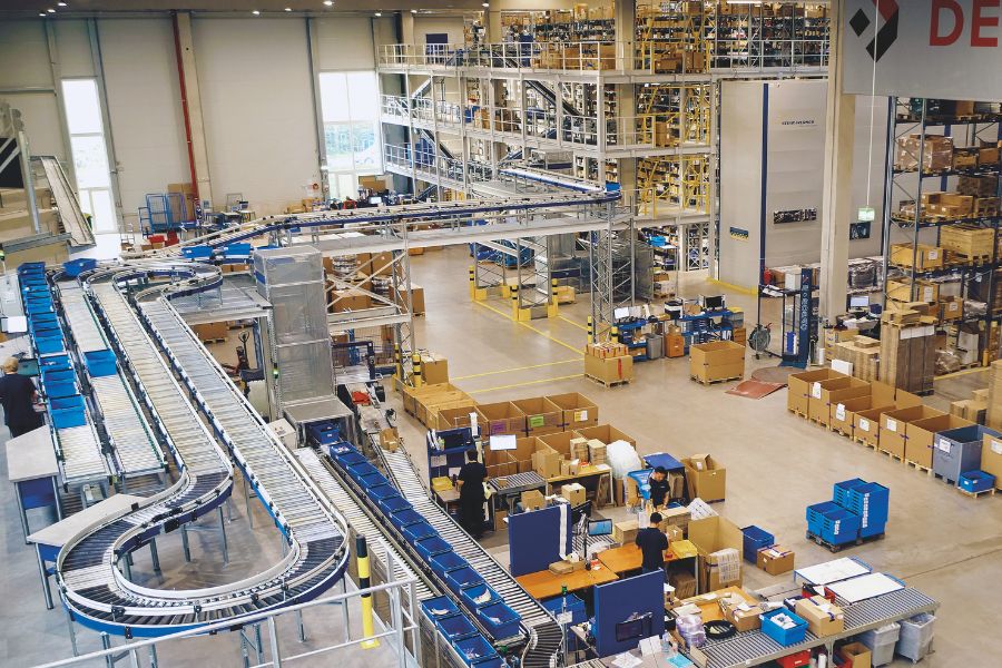 Ein weitläufiges Lager mit zahlreichen Förderbändern, die Pakete transportieren. Arbeiter arbeiten an verschiedenen Stationen auf der Etage und sortieren und verpacken Artikel. Im Hintergrund sind Regale voller Kisten und Waren sowie hohe Industrieanlagen zu sehen.