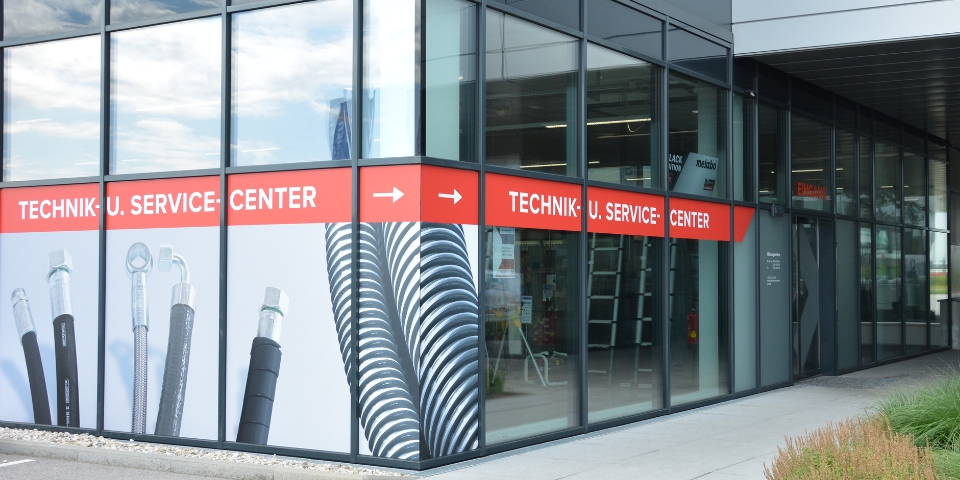 Das Bild zeigt die Außenfassade eines modernen Glasgebäudes mit einem roten Beklebung mit der Aufschrift „TECHNIK U. SERVICE CENTER“. Große Abbildungen von Schläuchen, die das technische Sortiment veranschaulichen, sind an den Fenstern zu sehen, und Pfeile weisen zum Eingang.