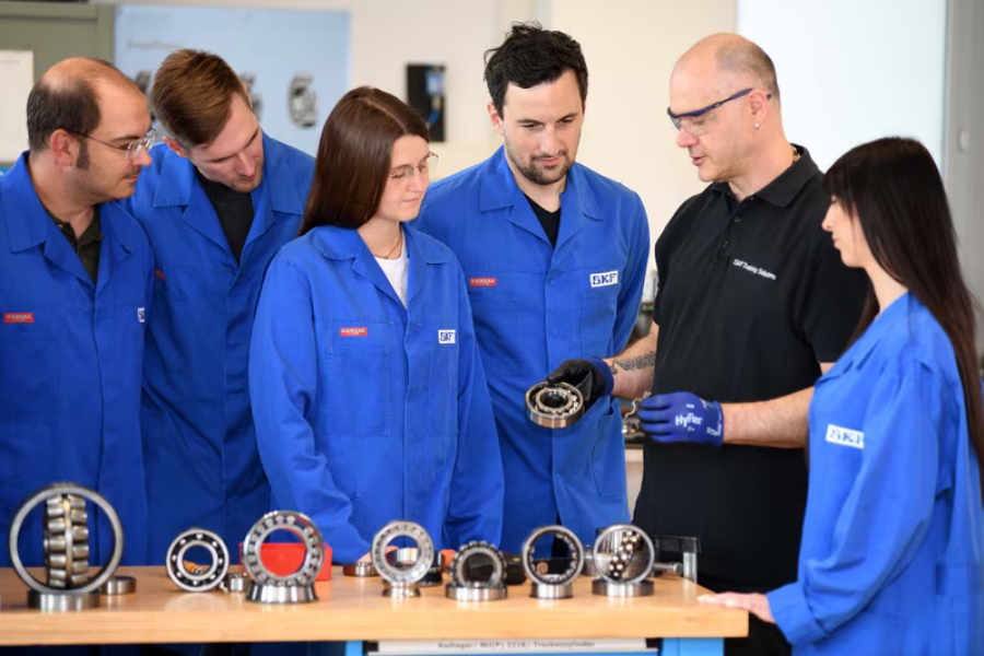 Eine Gruppe von Menschen in blauen Uniformen steht um einen Tisch mit verschiedenen Modellen mechanischer Lager. Eine Person in Schwarz mit Handschuhen erklärt der Gruppe etwas. Die Szenerie zeigt einen technischen Schulungsbereich.