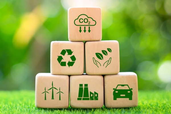 Holzklötze mit grünen Umweltsymbolen sind pyramidenförmig auf Gras gestapelt. Zu den Symbolen gehören CO2-Reduktion, Recycling, Blätter und Hände, Windräder, Industriegebäude und ein Elektroauto. Unscharfer grüner Hintergrund.