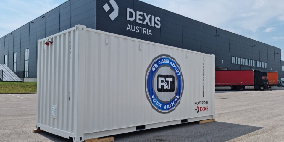 Weißer Frachtcontainer mit der Aufschrift "POWERED BY DEXIS" steht vor dem modernen DEXIS Austria Headquarter in Pasching. Im Hintergrund sind ein roter Lastwagen und ein schwarzer LKW-Anhänger zu sehen.  