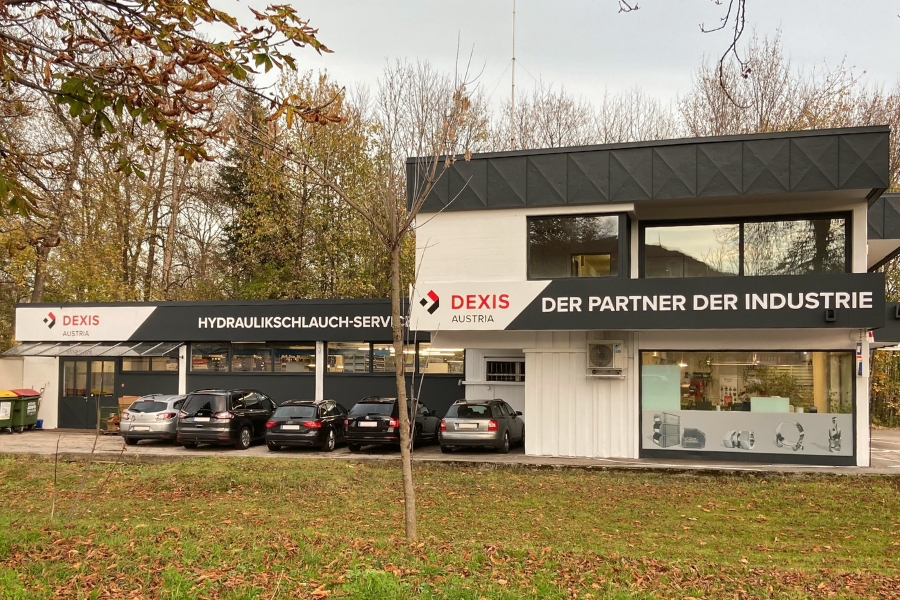 Firmengebäude in Villach mit der Aufschrift „DEXIS Austria“, „HYDRAULIKSCHLAUCH-SERVICE“ und „DER PARTNER DER INDUSTRIE“. Fünf Autos parken vor dem Gebäude. Die Bäume rundherum vermitteln eine herbstliche Stimmung.