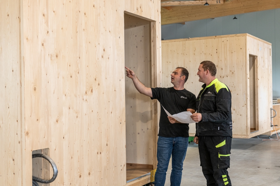 Zwei Männer in Arbeitskleidung besprechen im Innenbereich eine große Holzkonstruktion. Einer von ihnen hält Papiere in der Hand. Sie zeigen auf die Konstruktion, während sie sich auf die Planung konzentrieren.