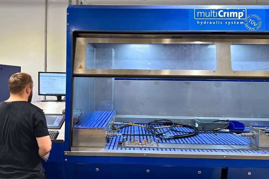 Ein Arbeiter mit schwarzem T-Shirt steht mit dem Rücken zur Kamera neben einer großen Hydraulikschlauchprüfstation mit der Aufschrift „multiCrimp hydraulic system“. Neben ihm befinden sich Hydraulikschläuche, Armaturen und Werkzeuge auf einem blauen Gittertisch. Die Umgebung zeigt die Prüfung von Hydraulikkomponenten.