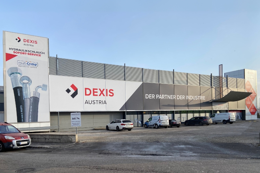 Firmengebäude in Brunn. Die Außenansicht zeigt ein Gewerbegebäude mit deutlichen Aufschriften für „DEXIS Austria“ und „DER PARTNER DER INDUSTRIE“. Mehrere geparkte Fahrzeuge säumen die Vorderseite des Gebäudes unter einem strahlend klaren Himmel.