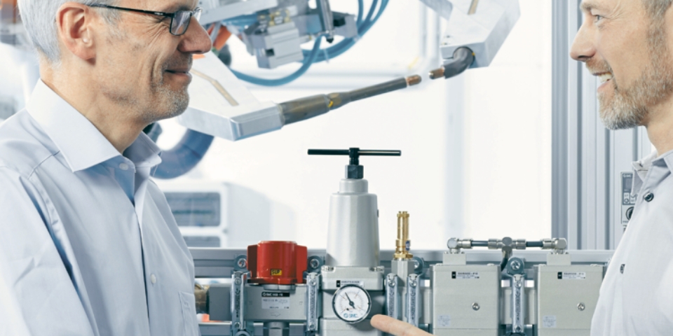 Zwei Männer stehen neben Industrieanlagen und unterhalten sich über die Maschinen. Im Hintergrund sind metallische Rohre, Messgeräte und Ventile zu sehen.