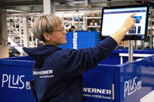 Eine Mitarbeiterin mit Brille und dunkelblauem Kapuzenpullover interagiert mit einem Touchscreen-Monitor, welcher auf dem RFID Gate befestigt ist, in einem Lager, umgeben von blauen Behältern mit der Aufschrift „RFID Gate“ und anderen Industriegeräten.