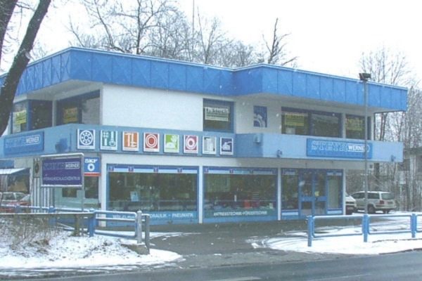 Steyr-Werner Standort in Villach 1994. Zweistöckiges blau-weiße Gebäude, mit bunten Schildern wo die Sortimente abgebildet sind. Schnee bedeckt den Boden und die Bäume, in der Nähe parken ein paar Autos unter dem bedeckten Himmel.