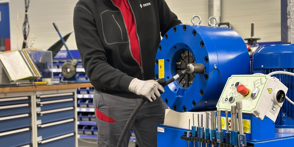 Eine Person mit Handschuhen bedient in einer Werkstatt eine blaue Hydraulikschlauchpressmaschine. Maschine und Werkzeuge sind gut sichtbar, im Hintergrund sind Regale mit Geräten zu sehen.