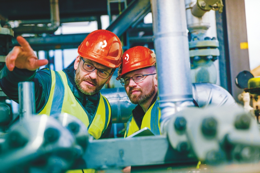 Zwei Mitarbeiter mit roten Schutzhelmen und Warnwesten sind auf einem Industriegelände. Ein Mitarbeiter zeigt nach vorne, während der andere in die gleiche Richtung blickt. Beide wirken konzentriert.