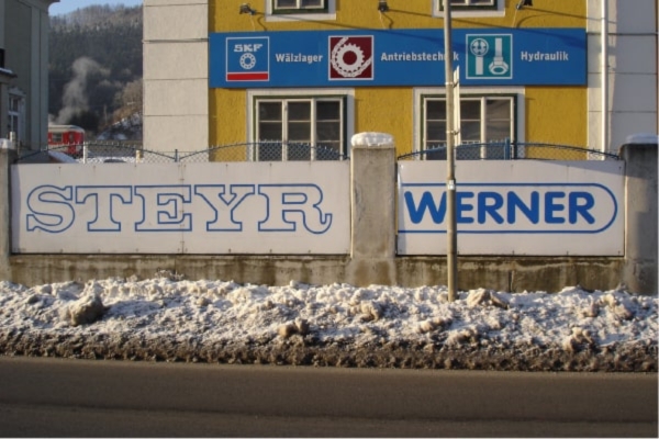 Eine Straßenansicht eines Gebäudes mit dem Schild „STEYR WERNER“. Die gelbe Fassade wird von einem verschneiten Gehweg und einem Maschendrahtzaun eingerahmt. Bäume und Hügel sieht man im Hintergrund..