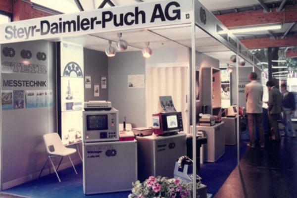 Der Messestand der „Steyr-Daimler-Puch AG“, präsentiert ein Sortiment an technischen Messgeräten. Poster säumen die Wände, während sich zahlreiche Besucher in der Nähe versammeln und die Ausstellung bewundern. Topfblumen sind ebenfalls zu sehen.