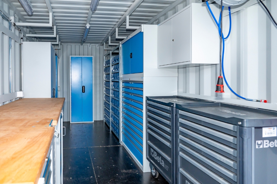Innenansicht eines Containers, der als Lagercontainer dient. Auf der linken Seite befindet sich eine lange Werkbank aus Holz, auf der rechten Seite mehrere graue Werkstattschränke und Schubladensysteme. Ganz hinten ist blaue Tür von einem weiteren hohen Werkstattschrank sichtbar. An der Decke verlaufen Kabel und Leitungen mit Licht.  