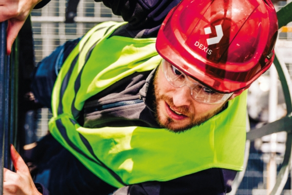Ein Bauarbeiter trägt einen roten Schutzhelm mit Logo, eine Schutzbrille und eine leuchtend gelbe Warnweste und blickt nach oben. Im Hintergrund sind Metallkonstruktionen und Gerüste zu sehen, die auf eine Baustelle hindeuten.