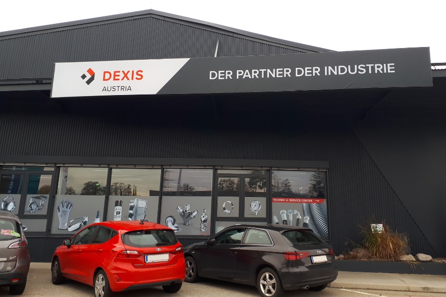 Außenansicht der DEXIS Austria Filiale in Graz mit dem Banner „Der Partner der Industrie“. Zwei Autos parken vor dem Gebäude. Auf der Fensterfolierung wird das technisches Sortiment präsentiert.