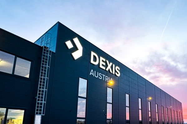Das modernes Firmengebäude von DEXIS Austria in Pasching.  Man sieht große Fenstern und das „DEXIS AUSTRIA“ Logo ragt stolz empor. Das elegante, dunkle Äußere ergänzt den Farbverlauf des Himmels in Blau und Violett und erinnert an Sonnenauf- oder -untergang.