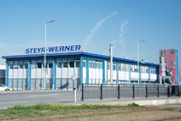 Ein blau-weißes Industriegebäude, mit der Aufschrift „STEYR-WERNER“ auf dem Dach, steht an einer Straße. Im Vordergrund ist ein Zaun zu sehen, rechts davon ein großes vertikales Banner mit unscharfen Motiven unter strahlend blauem Himmel.