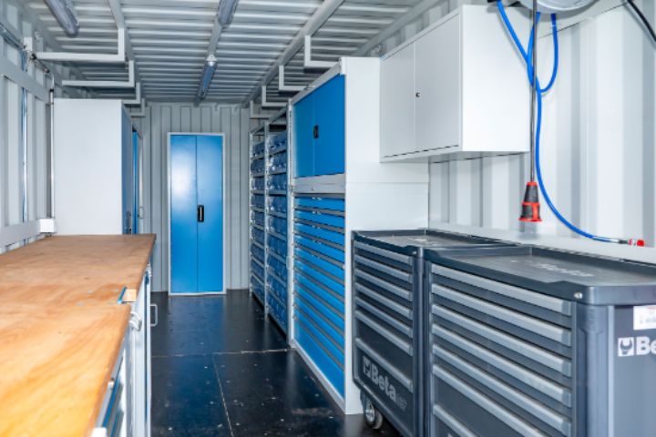 Innenansicht eines Containers, der als Lagercontainer dient. Auf der linken Seite befindet sich eine lange Werkbank aus Holz, auf der rechten Seite mehrere graue Werkstattschränke und Schubladensysteme. Ganz hinten ist blaue Tür von einem weiteren hohen Werkstattschrank sichtbar. An der Decke verlaufen Kabel und Leitungen mit Licht.  