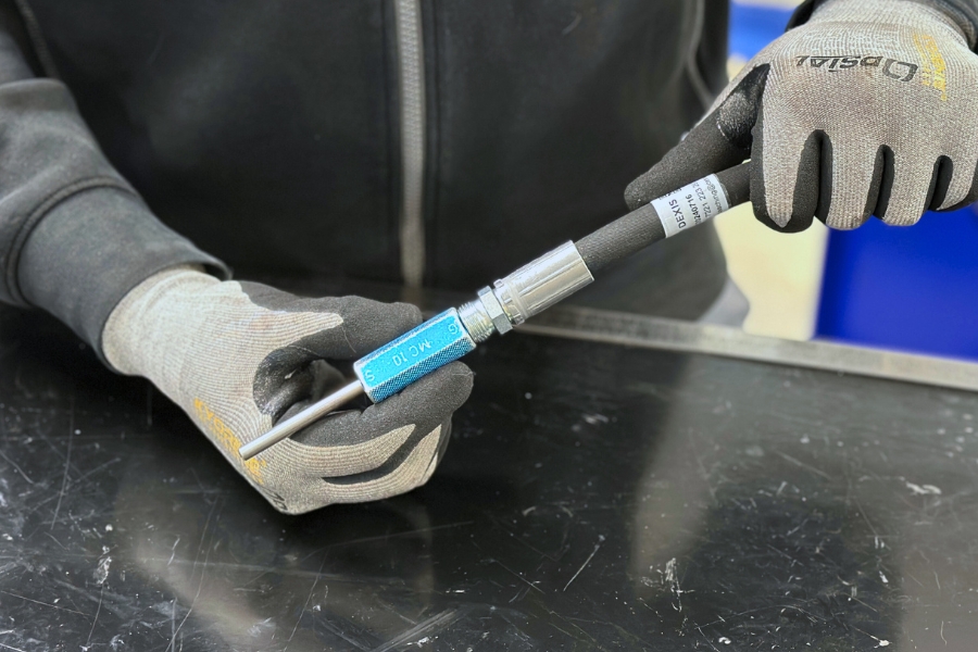 Eine Person mit grauen OPSIAL Handschuhen hält ein Prüfdorn mit einer blauen Kennzeichnung in den Händen und einen Hydraulikschlauch. Der Prüfdorn steckt mit einer Seite in dem Hydraulikschlauch. Die Szene spielt sich auf einer schwarzen Werkbank ab.