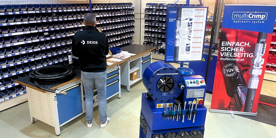 Eine Mitarbeiter in einer schwarzen DEXIS-Jacke steht in einer organisierten Werkstatt und konzentriert sich auf Gegenstände auf einem Tisch. An der Wand stehen Regale voller Teile. Im Vordergrund steht eine blaue Hydraulikmaschine, daneben hängt ein multiCrimp-Banner.