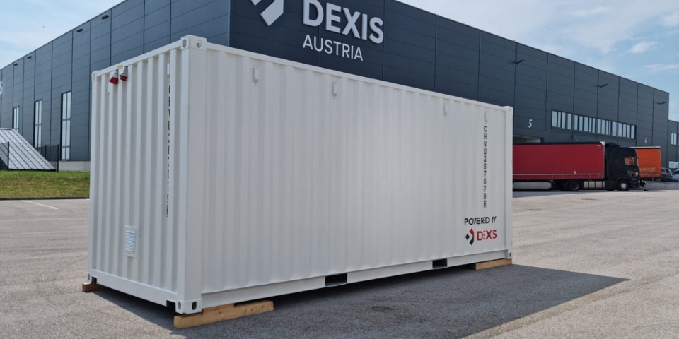 Weißer Frachtcontainer mit der Aufschrift "POWERED BY DEXIS" steht vor dem modernen DEXIS Austria Headquarter in Pasching. Im Hintergrund sind ein roter Lastwagen und ein schwarzer LKW-Anhänger zu sehen.  