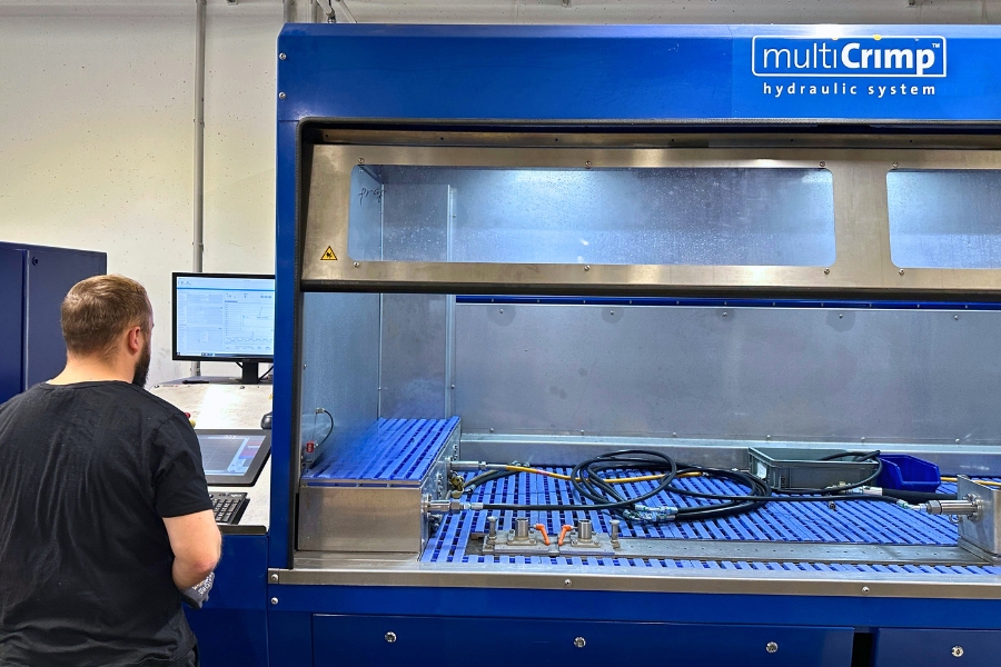 Ein Arbeiter mit schwarzem T-Shirt steht mit dem Rücken zur Kamera neben einer großen Hydraulikschlauchprüfstation mit der Aufschrift „multiCrimp hydraulic system“. Neben ihm befinden sich Hydraulikschläuche, Armaturen und Werkzeuge auf einem blauen Gittertisch. Die Umgebung zeigt die Prüfung von Hydraulikkomponenten.