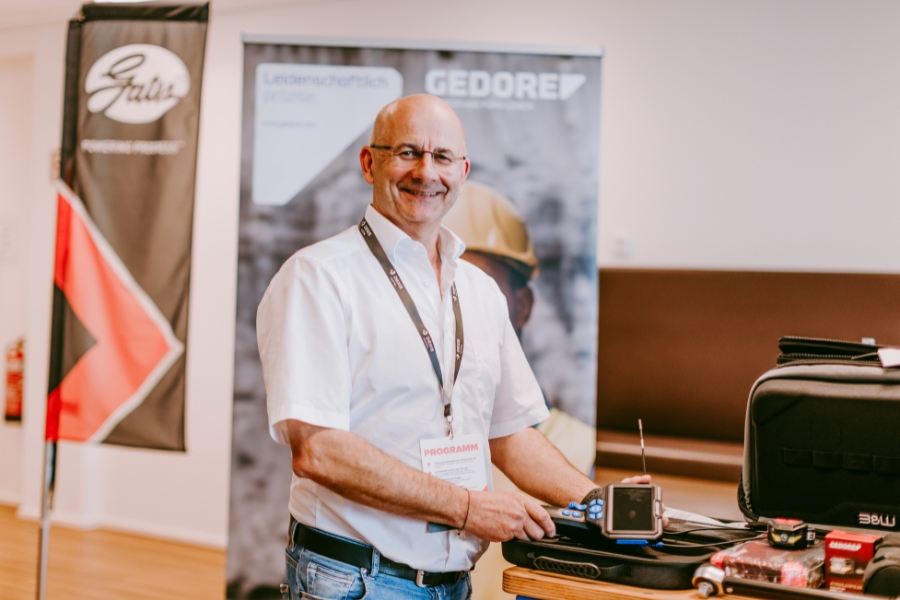 Ein Mann im weißen Hemd steht lächelnd vor einem Stand mit „Gedore“-Roll-up. Er hält ein Werkzeug in der Hand, während auf dem Tisch neben ihm weitere Werkzeuge ausgestellt sind. Fahnen und Banner schmücken den Hintergrund.