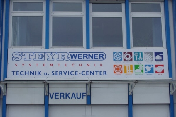 Das Bild zeigt die Außenansicht eines Gebäudes mit einem Schild „Steyr Werner Systemtechnik“. Bunte Symbole repräsentieren verschiedene Dienstleistungen, während ein blaues „Verkauf“-Schild die Szenerie abrundet. Das Gebäude ist mit mehreren Fenstern und blauen Zierleisten geschmückt.