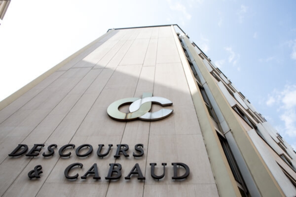 Untersicht eines hohen Gebäudes mit dem Schriftzug „Descours & Cabaud“ und einem Logo an der Fassade. Das helle Gebäude steht vor einem leicht bewölkten Himmel.