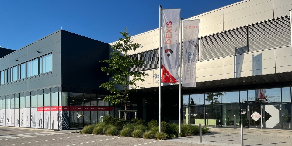 Modernes Industriegebäude mit grauer Fassade. Davor ist eine Grünflächen mit Baum angelegt, wo auch zwei Fahnen stehen mit "DEXIS Austria" und "multiCrimp".  Glaswände geben den Blick auf das Technik und Service Center sowie den Eingangsbereich des Bürogebäudes frei. Es ist strahlend blauer Himmel zu sehen.