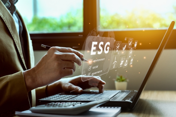 Eine Person im Anzug bedient einen Laptop. Über der Tastatur ist eine digitale Einblendung mit Symbolen und den Buchstaben „ESG“ (Environment, Social, Governance) zu sehen. Im Hintergrund fällt Sonnenlicht durch ein Fenster.