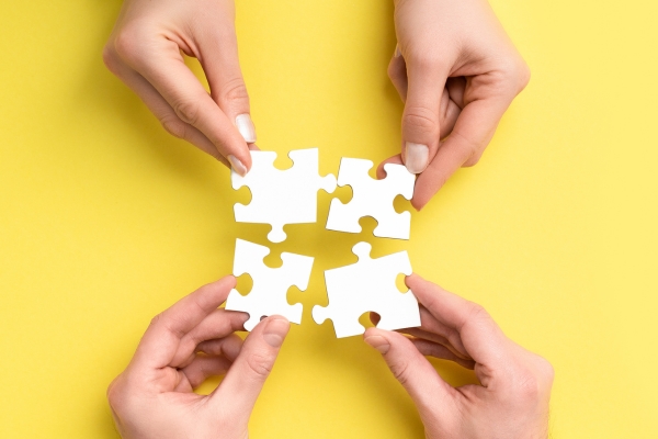 Zwei Handpaare mit Puzzleteilen in je einer Hand. Beide versuchen die Puzzleteile mit einander zu verbinden. Der Hintergrund ist ein kräftiges gelb.