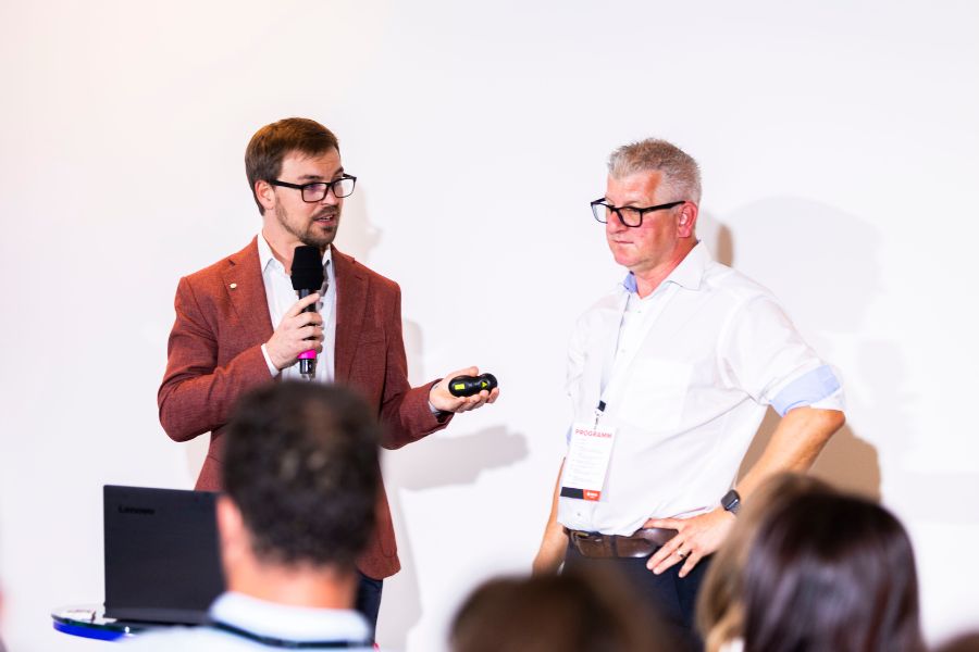 In einem Konferenzraum sitzen Menschen vor einer Leinwand, auf der zwei Moderatoren von DEXIS Austria sprechen. Das aufmerksame Publikum verfolgt die auf die Leinwand projizierten Folien, die das Einsparungspotenziale mit Hilfe von DEXIS präsentieren. Der Raum verfügt über einen Deckenprojektor und weiße Wände.