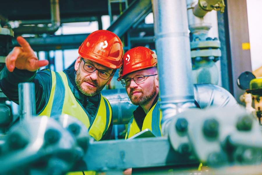 Zwei Mitarbeiter mit roten Schutzhelmen und Warnwesten sind auf einem Industriegelände. Ein Mitarbeiter zeigt nach vorne, während der andere in die gleiche Richtung blickt. Beide wirken konzentriert.