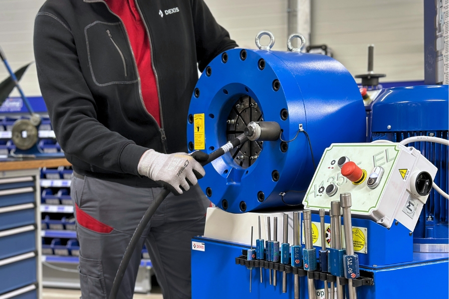 Ein Arbeiter in schwarzer und roter Arbeitskleidung mit Handschuhen bedient eine blaue Schlauchpresse, um einen Hydraulikschlauch zu verpressen. Die Maschine hat eine runde Öffnung mit mehreren Spannbacken, in die der Schlauch eingeführt wird. Daneben befindet sich ein Bedienfeld mit einem Not-Aus-Knopf und anderen Steuerelementen. Im Hintergrund sind eine Werkbank und Werkzeuge zu sehen, was auf eine Werkstattumgebung hinweist.