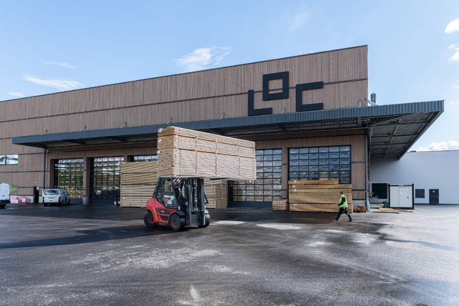 Ein Gabelstapler manövriert gekonnt einen großen Stapel Holzbretter vor einer modernen Lagerhalle mit den Buchstaben „LOC“. Ein Mitarbeiter in Warnweste überwacht den Vorgang. Im Hintergrund erkennt man den klaren und sonnigen Himmel.