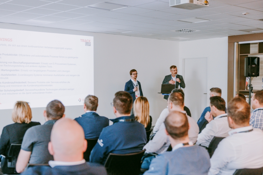 In einem Konferenzraum sitzen Menschen vor einer Leinwand, auf der zwei Moderatoren von DEXIS Austria sprechen. Das aufmerksame Publikum verfolgt die auf die Leinwand projizierten Folien, die das Einsparungspotenziale mit Hilfe von DEXIS präsentieren. Der Raum verfügt über einen Deckenprojektor und weiße Wände.