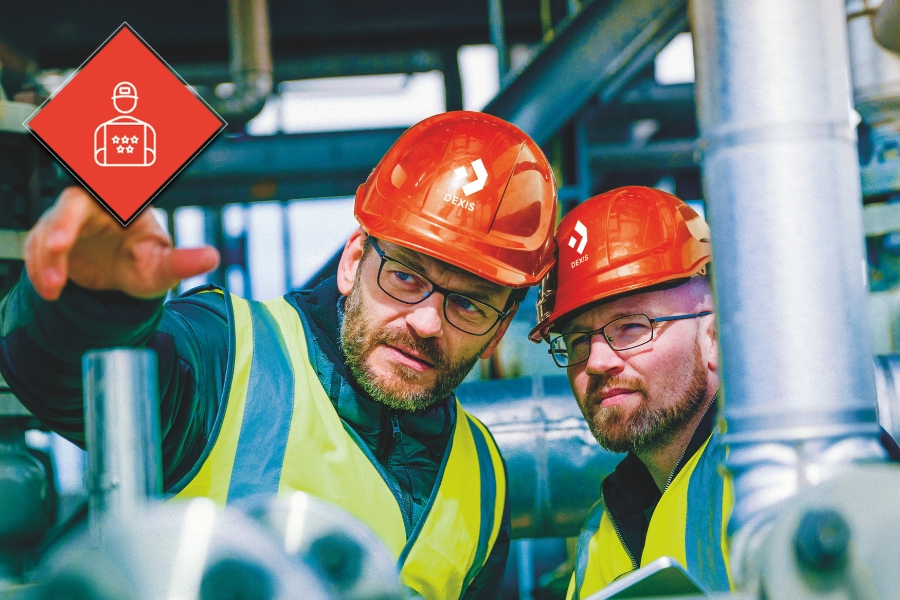 Zwei Mitarbeiter mit roten Schutzhelmen und Warnwesten sind auf einem Industriegelände. Ein Mitarbeiter zeigt nach vorne, während der andere in die gleiche Richtung blickt. Beide wirken konzentriert. In der oberen linken Ecke ist  eine rote Raute mit einem Symbol das einen Experten darstellt.
