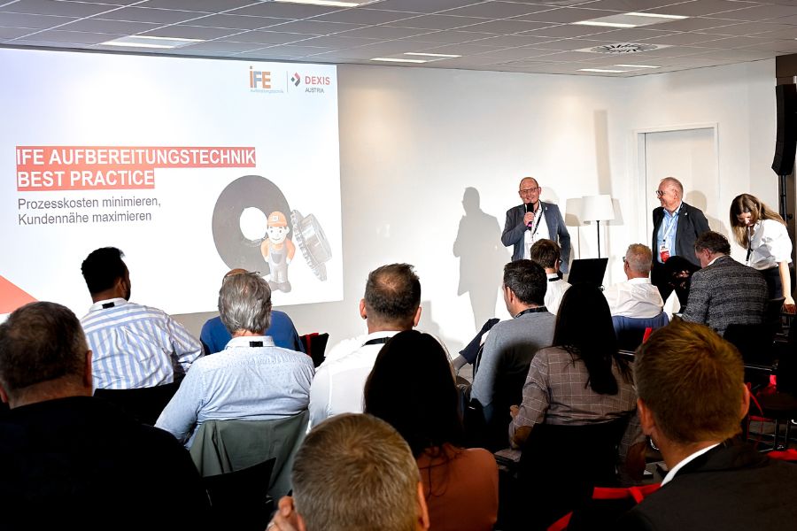 In einem Konferenzraum sitzen Menschen vor einer Leinwand, auf der zwei Moderatoren von DEXIS Austria sprechen. Das aufmerksame Publikum verfolgt die auf die Leinwand projizierten Folien, die das Einsparungspotenziale mit Hilfe von DEXIS präsentieren. Der Raum verfügt über einen Deckenprojektor und weiße Wände.