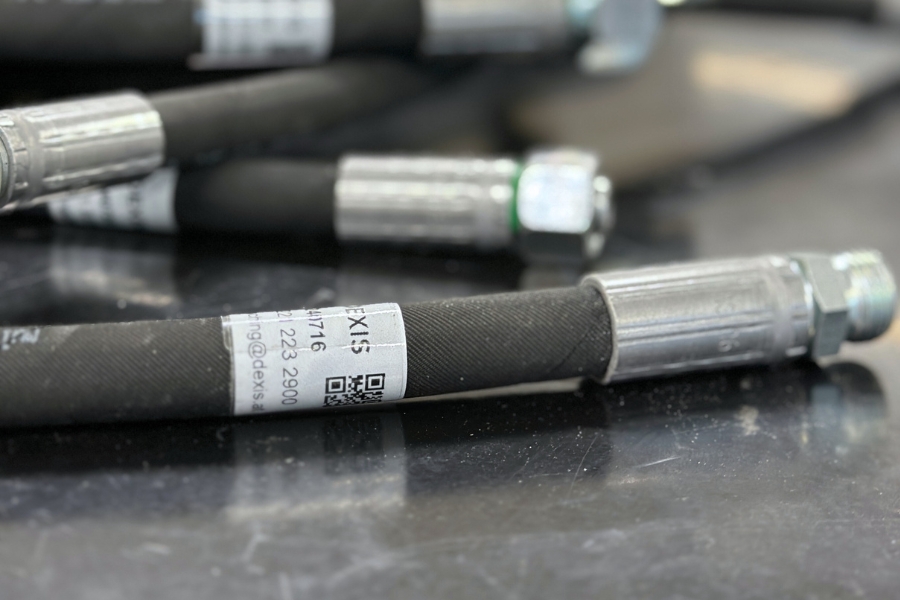 Nahaufnahme von Industrieschläuchen mit Metallanschlüssen. Ein Schlauch trägt ein Etikett mit der Aufschrift „EXIS“ und einen QR-Code. Die Schläuche sind schwarz mit einer reflektierenden Oberfläche und liegen auf einer dunklen, glänzenden Tischplatte.