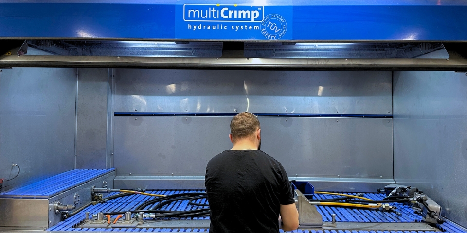 Ein Arbeiter mit schwarzem T-Shirt steht mit dem Rücken zur Kamera an einer großen Arbeitsstation mit der Aufschrift „multiCrimp hydraulic system“. Vor ihm befinden sich Hydraulikschläuche, Armaturen und Werkzeuge auf einem blauen Gittertisch. Die Umgebung zeigt die Prüfung von Hydraulikkomponenten.