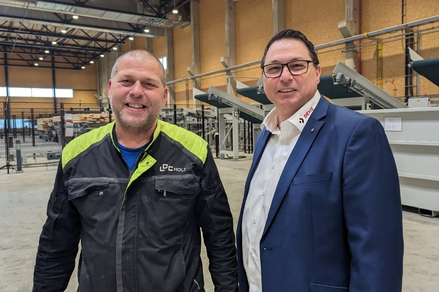 Zwei Männer stehen Seite an Seite in einer Industrieanlage. Der Mann links trägt eine schwarz-neongelbe Arbeitsjacke, der Mann rechts einen blauen Blazer und ein weißes Hemd. Beide lächeln selbstbewusst in die Kamera.