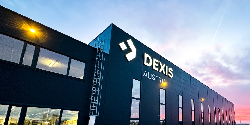 Das modernes Firmengebäude von DEXIS Austria in Pasching.  Man sieht große Fenstern und das „DEXIS AUSTRIA“ Logo ragt stolz empor. Das elegante, dunkle Äußere ergänzt den Farbverlauf des Himmels in Blau und Violett und erinnert an Sonnenauf- oder -untergang.